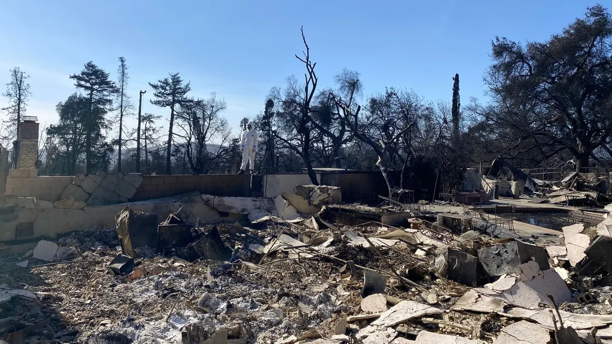 A person in a white protective suit stands in the ruins of a home destroyed by fire