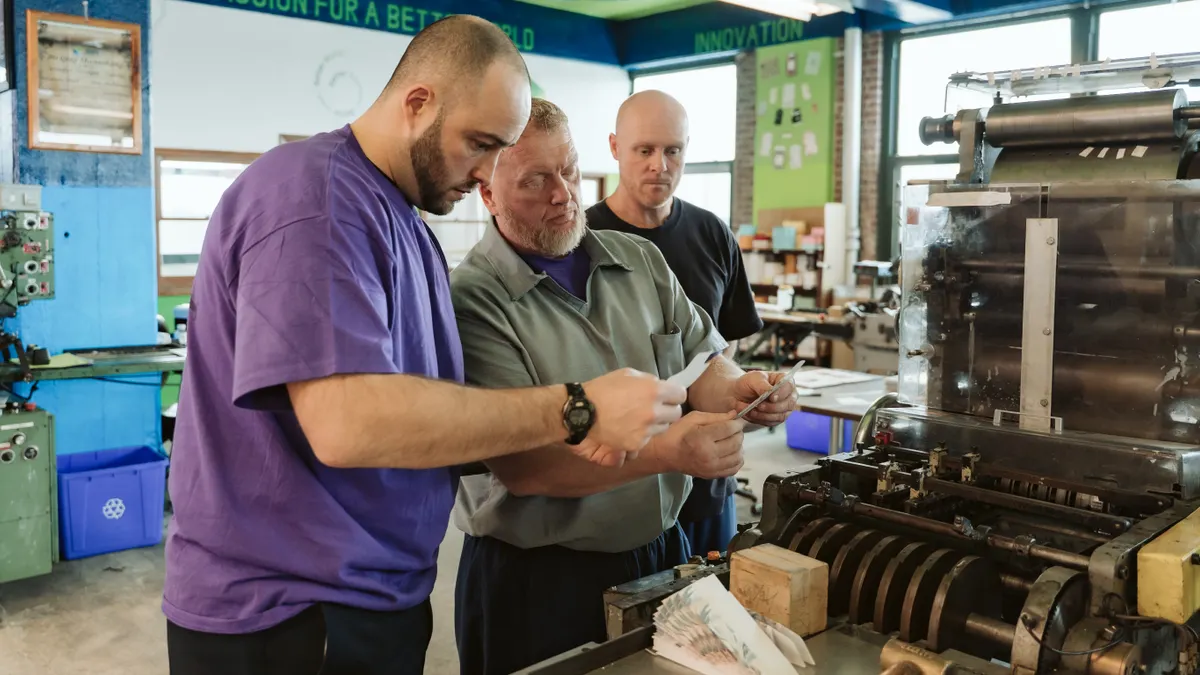 Three people examine envelopes while standing next to an industrial machine.