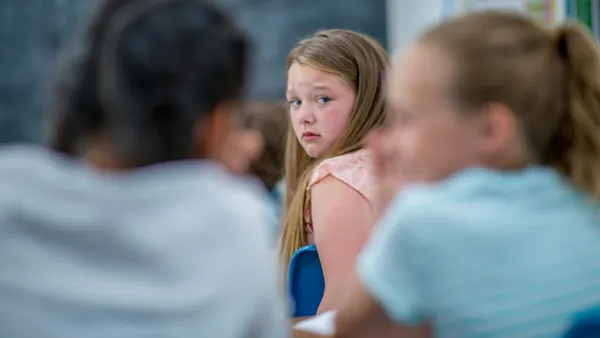 A student is sitting at a desk in a classroom and looking over their shoulder at two other students who have their backs to the camera.