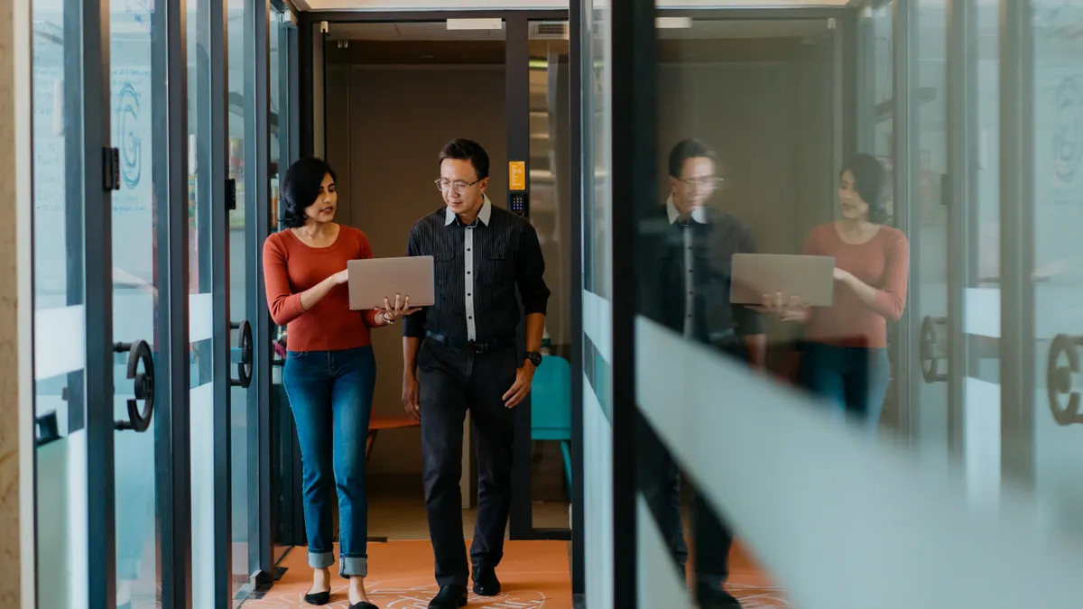 Two tech office workers walk down a hall looking at a laptop.