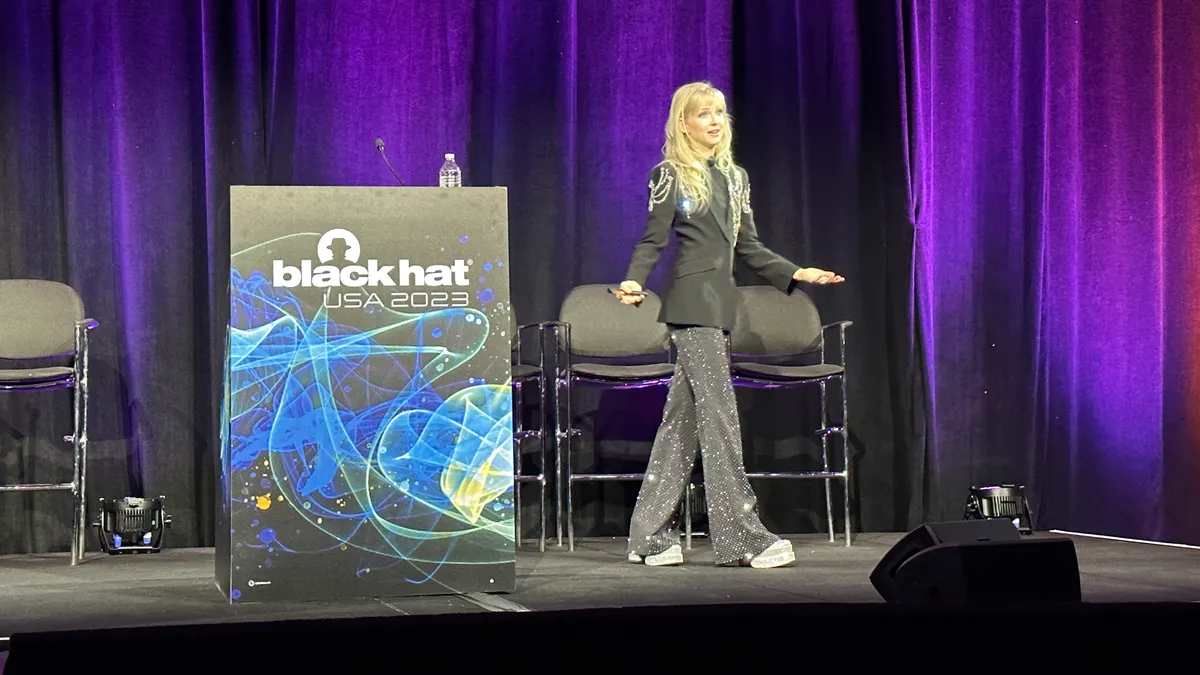 Woman speaking on stage in front of a purple curtain in a black blazer and sparkly pants. There is also a podium that says "Black Hat USA 2023."