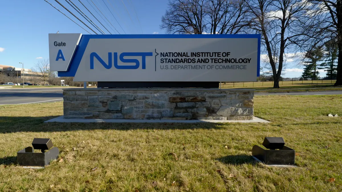 A large entrance sign that reads "Gate A, NIST, National Institute of Standards and Technology, U.S. Department of Commerce" is mounted on a rock base and surrounded by grass and trees. In the background to the left of the sign, there is a commercial building.